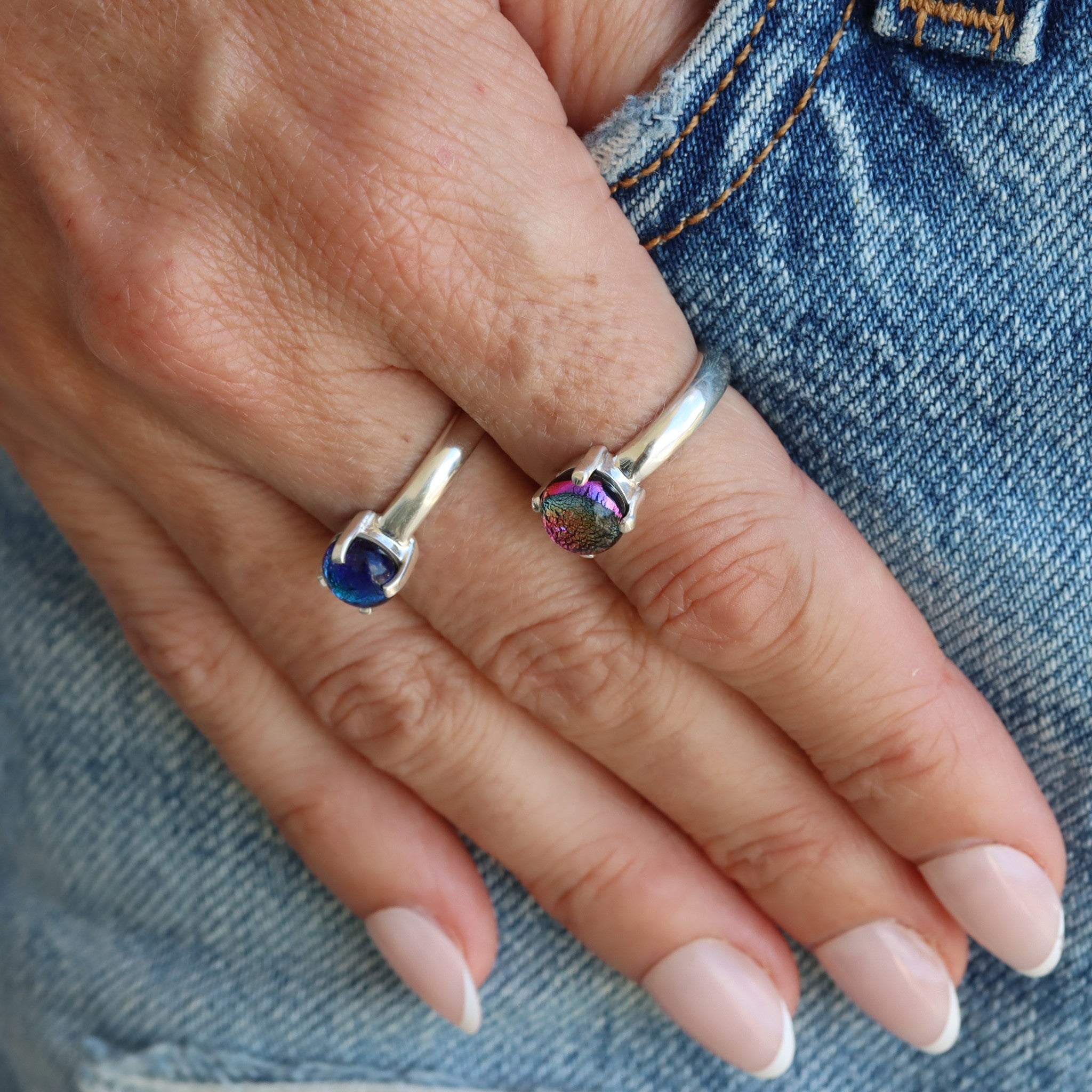 A hand wearing two sterling silver, small, round, claw-set dichroic glass gem rings. One in pink and the other in blue tones.