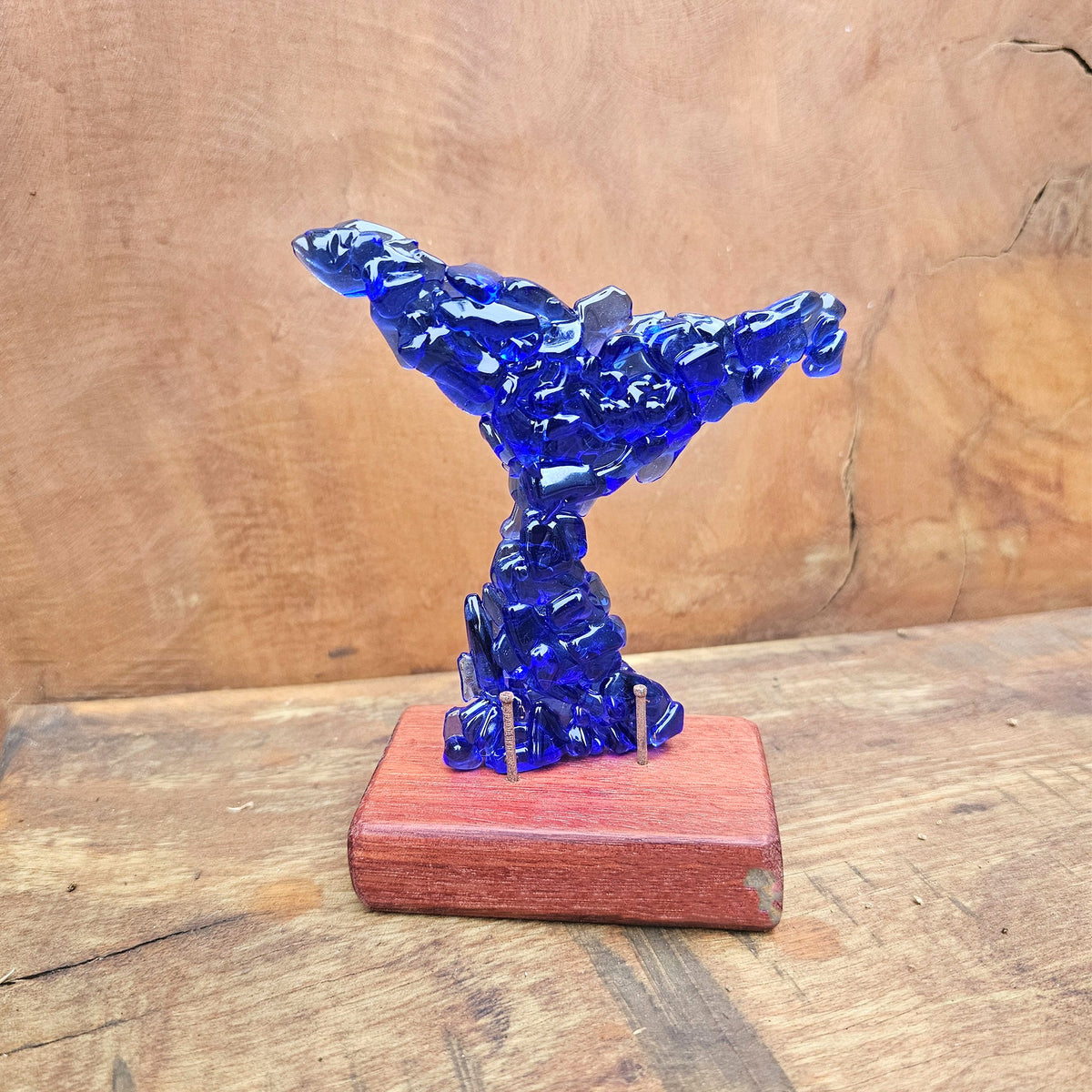 A dark ink blue translucent glass sculpture of a whale tail standing on a reddish-brown sanded and oiled rectangular wooden base, photographed against a rustic wood background.