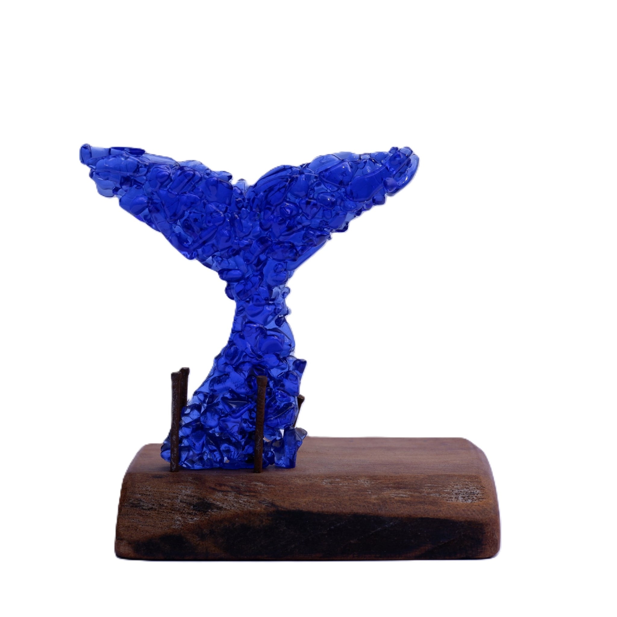 A translucent cobalt blue glass whale tail sculpture mounted on a rustic dark brown wooden base with rusted and aged nails, photographed against a solid white background.