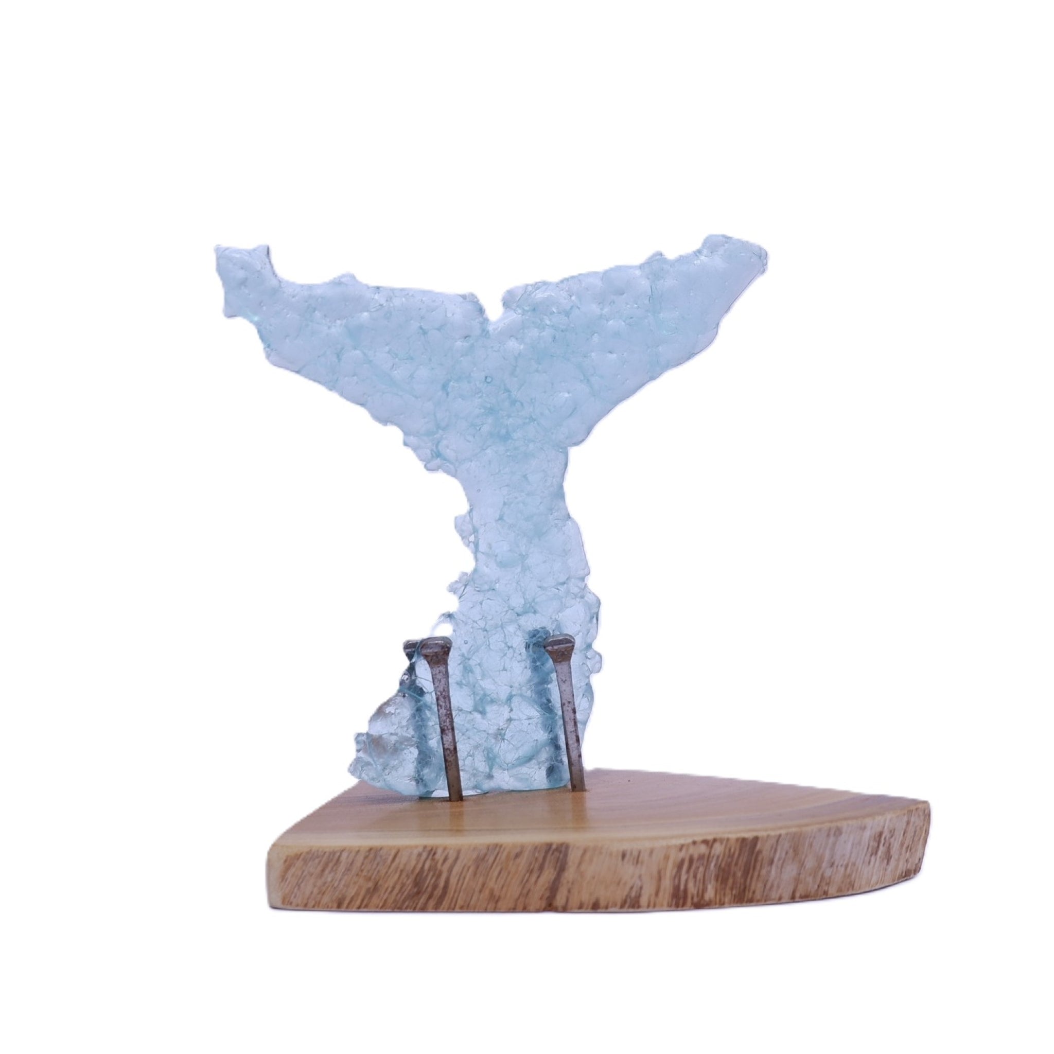 A decorative whale tail sculpture made of translucent light blue glass fragments, mounted on a natural wood slice base with 3 visible rusted and aged nails holding it in place, shown against a plain white background.