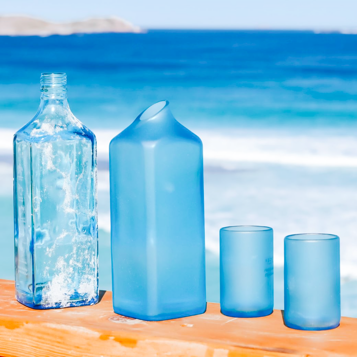 Three translucent blue drinking vessels on a wooden surface with a blurred ocean background: a tall square bottle with sand on the edge, a large rectangular drinking carafe, and two round mini glasses.