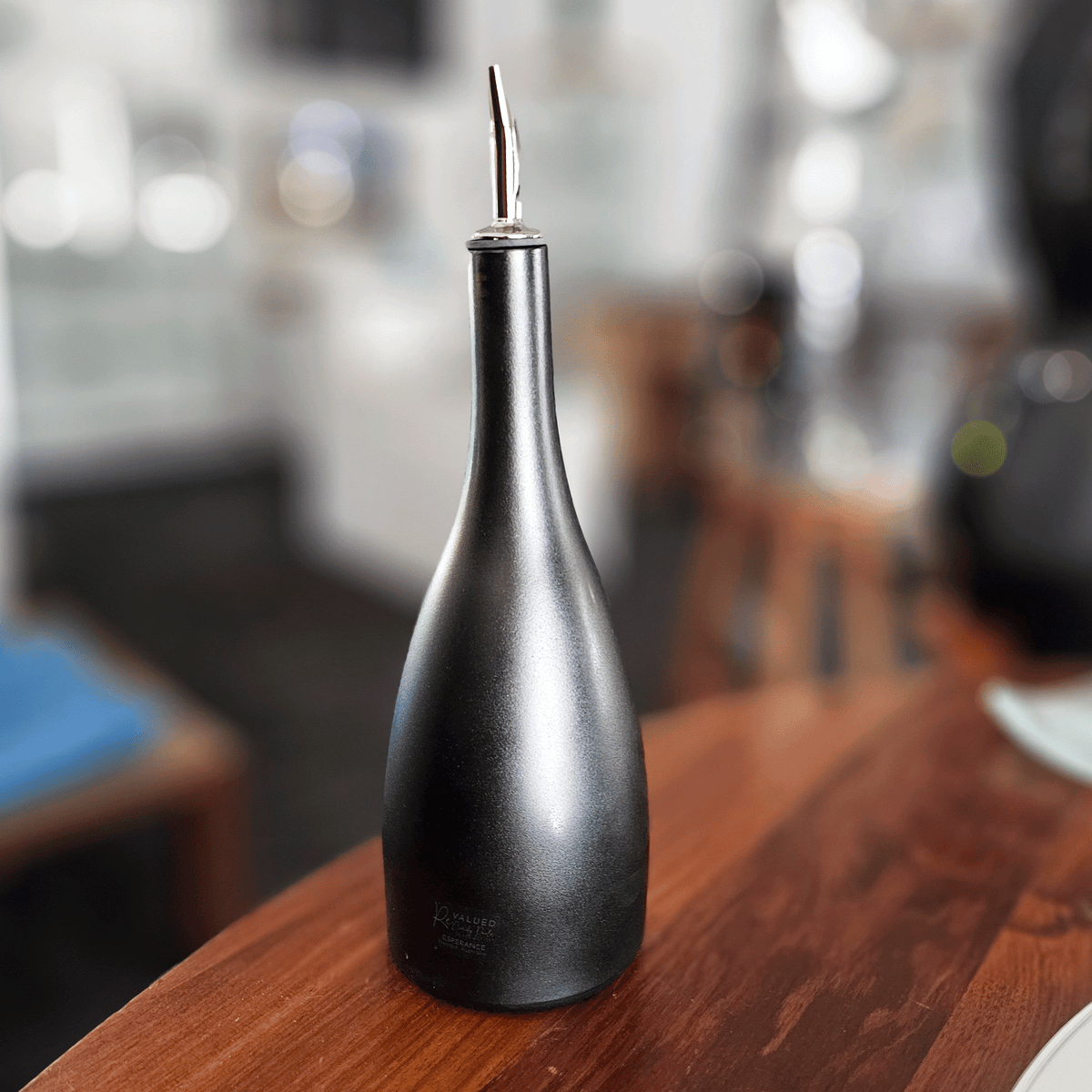 A matte black glass oil dispenser bottle with a stainless steel pour spout, standing on a wooden table in a blurred indoor setting.