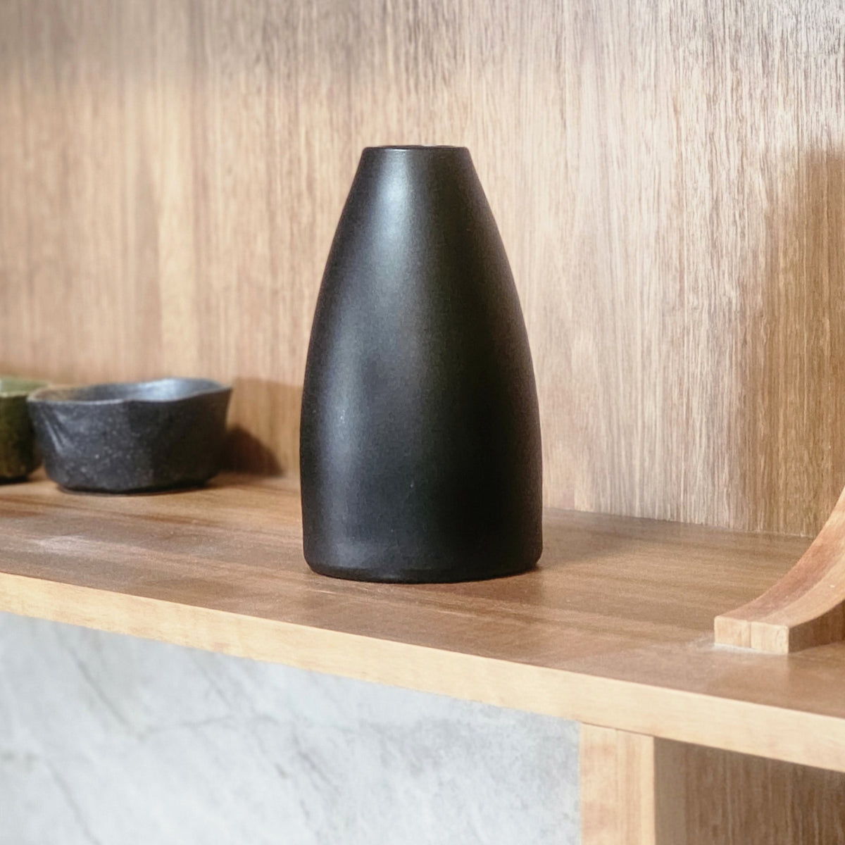 A matte obsidian hued glass vase with a tapered, rounded shape sits on a light wood shelf against a wood-paneled wall.