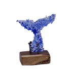 With blue hues and silver fragments this dichroic art glass whale tail sculpture is mounted on a sanded and oiled rectangular wooden base with visible aged and rusted nails. Photographed against a plain white background.