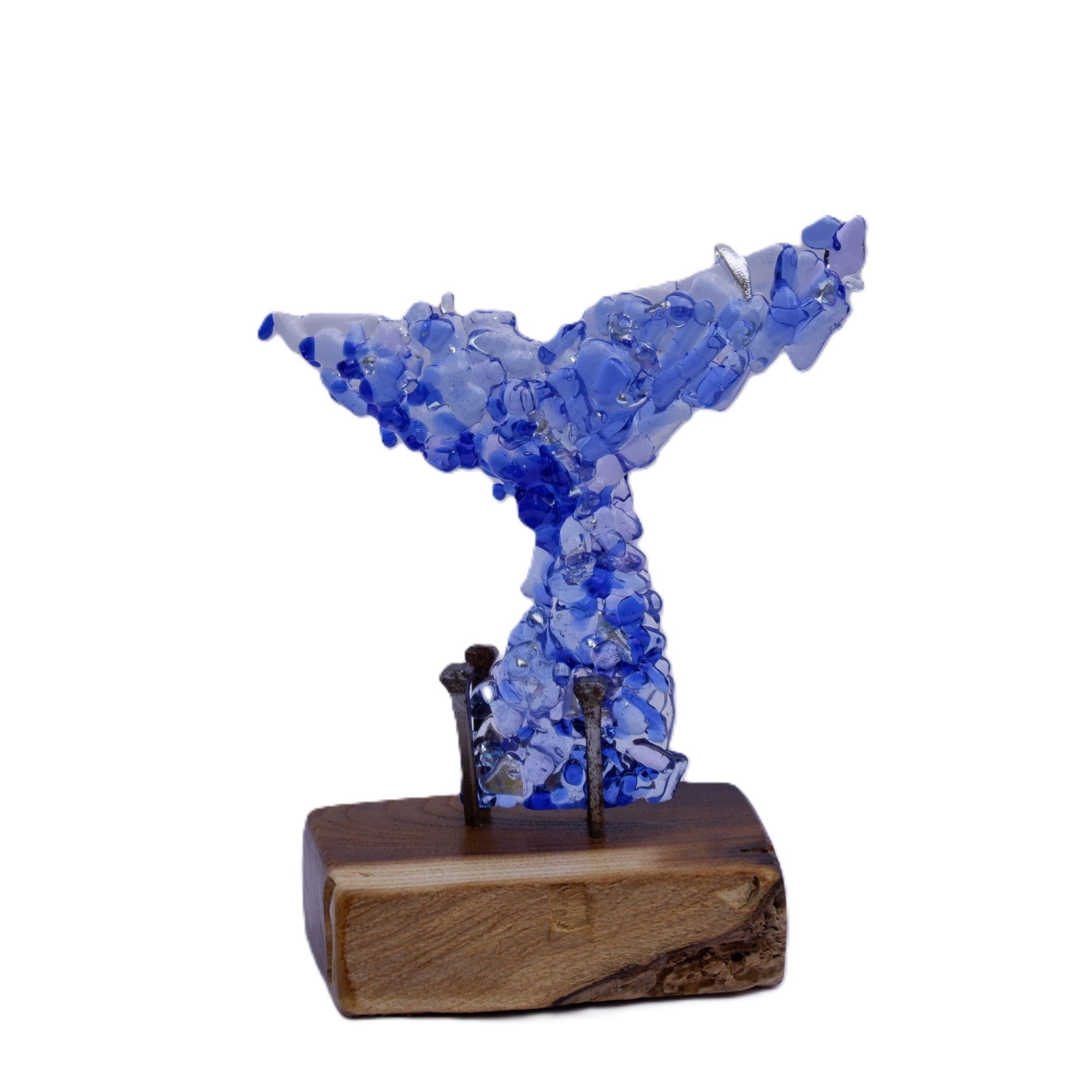With blue hues and silver fragments this dichroic art glass whale tail sculpture is mounted on a sanded and oiled rectangular wooden base with visible aged and rusted nails. Photographed against a plain white background.