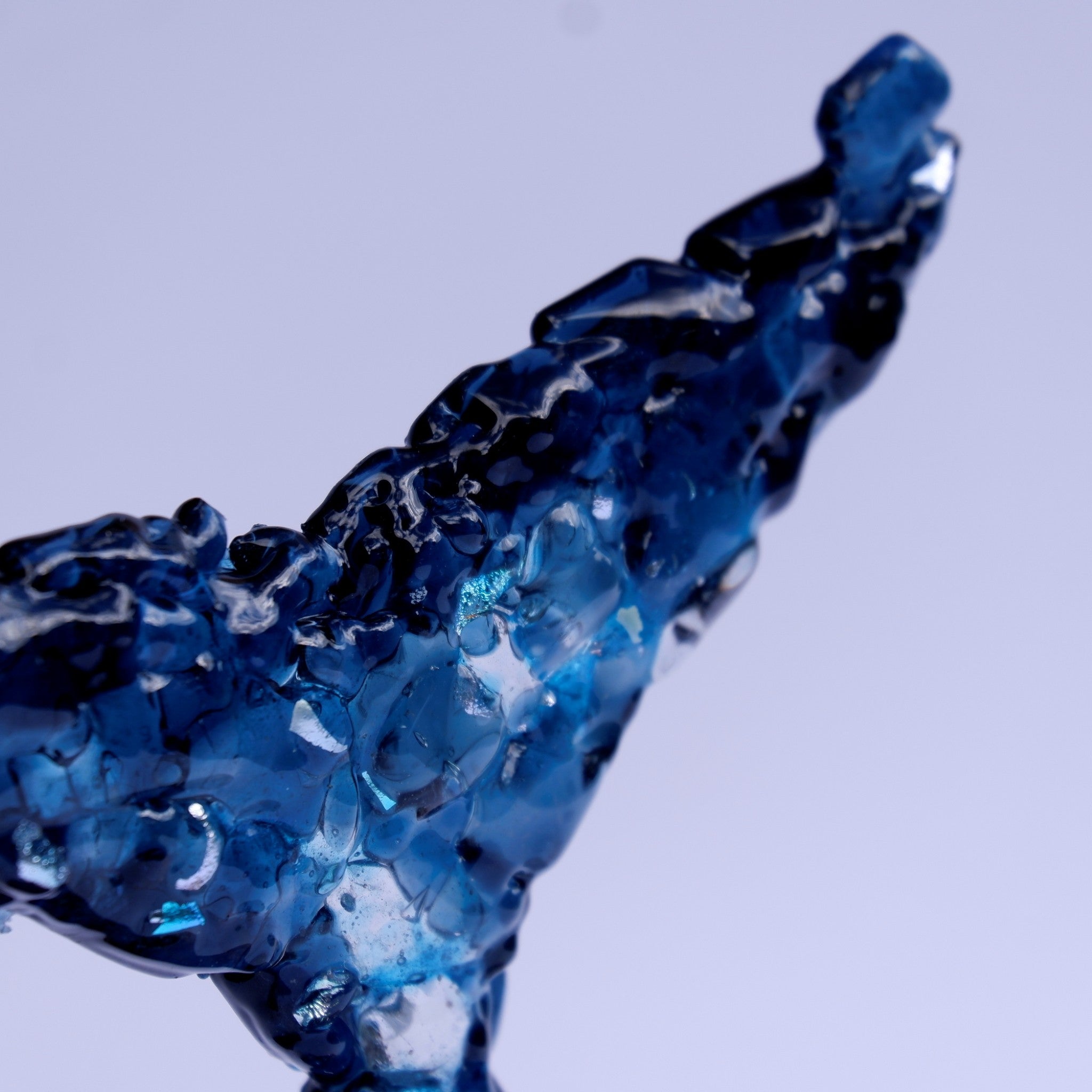 A close-up of a translucent blue dichroic and art glass whale tail sculpture, showing details of the textured fused glass fragments formingthe glossy refractile surface against a pale lavender toned background.