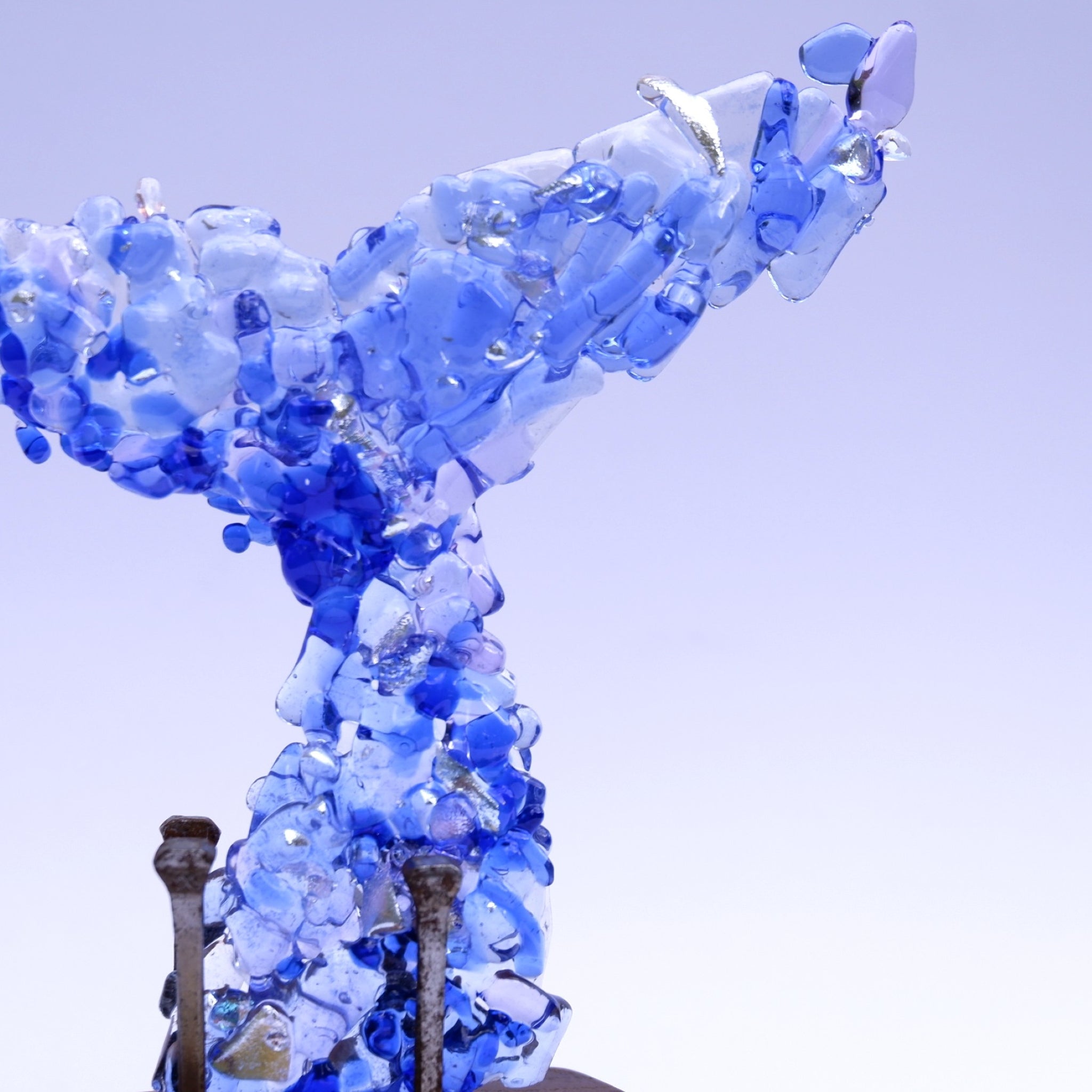 A close-up of a translucent blue, silver and ocean inspired glass whale tail sculpture made from fragments or irregular art & dichroic glass pieces.  Visible are a few rusted & aged metal nails visible at the base, against a pale lavender hued background.