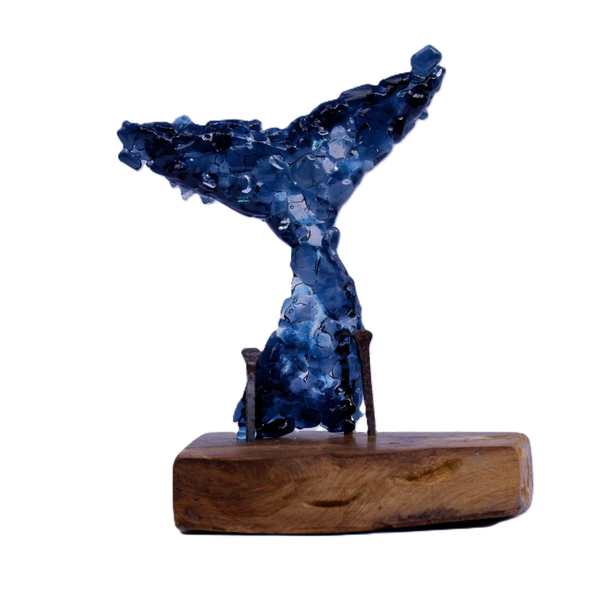 A dark blue and teal hued whale tail sculpture made of dichroic and art glass fragments, mounted on a sanded and oiled natural brown wooden base with two small nails, set against a plain white background.