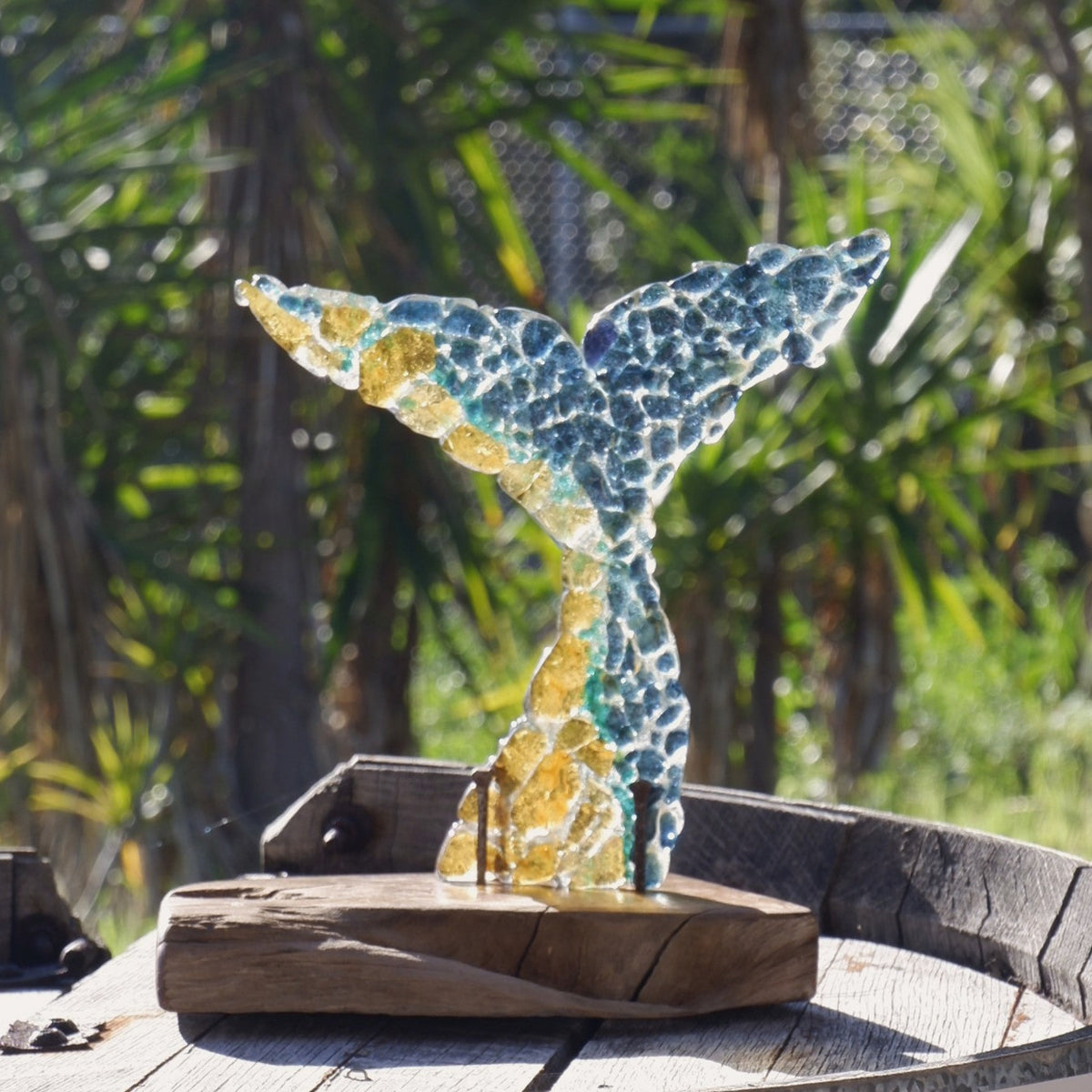 A fused art glass whale tail sculpture made of glass fragments in blue, green, and yellow, mounted on a rustic Australian Bush timber base outdoors on a weathered wooden table with trees in the background.