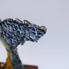 Close-up of a glossy, translucent, textured fused art glass whale tail fluke in dark blue and gold tones, set against a plain light gray background.