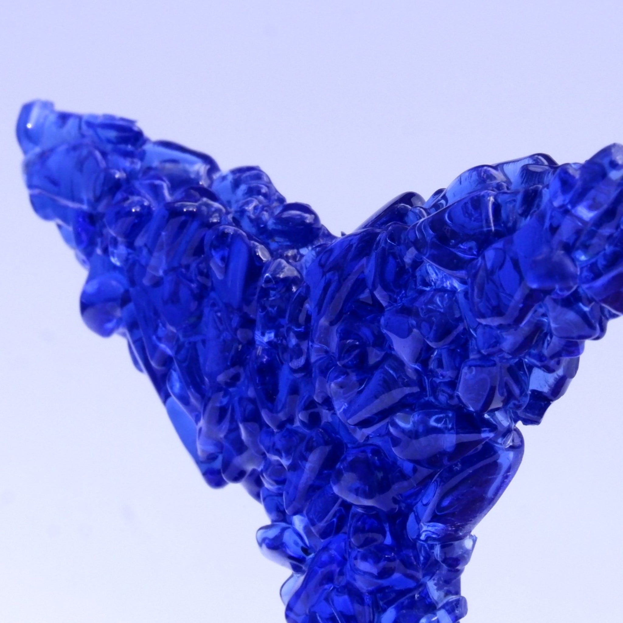 A close-up of a translucent cobalt blue glass whale tail flukes, a decorative sculpture of fused glass fragments, set against a cool pale lavender background.