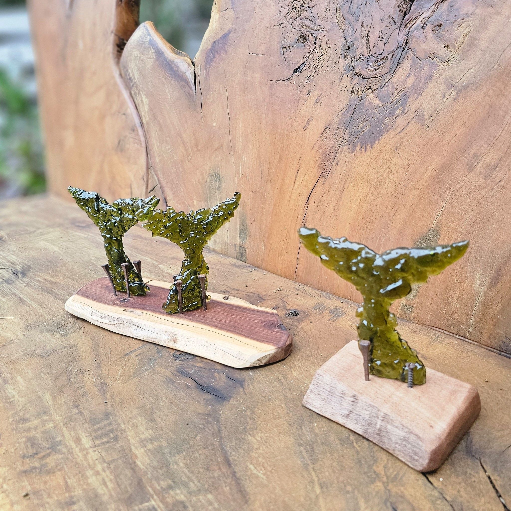 Two decorative whale tail sculptures made from kelp-olive-green glass fragments, mounted on natural wood bases, displayed on a rustic wooden surface.