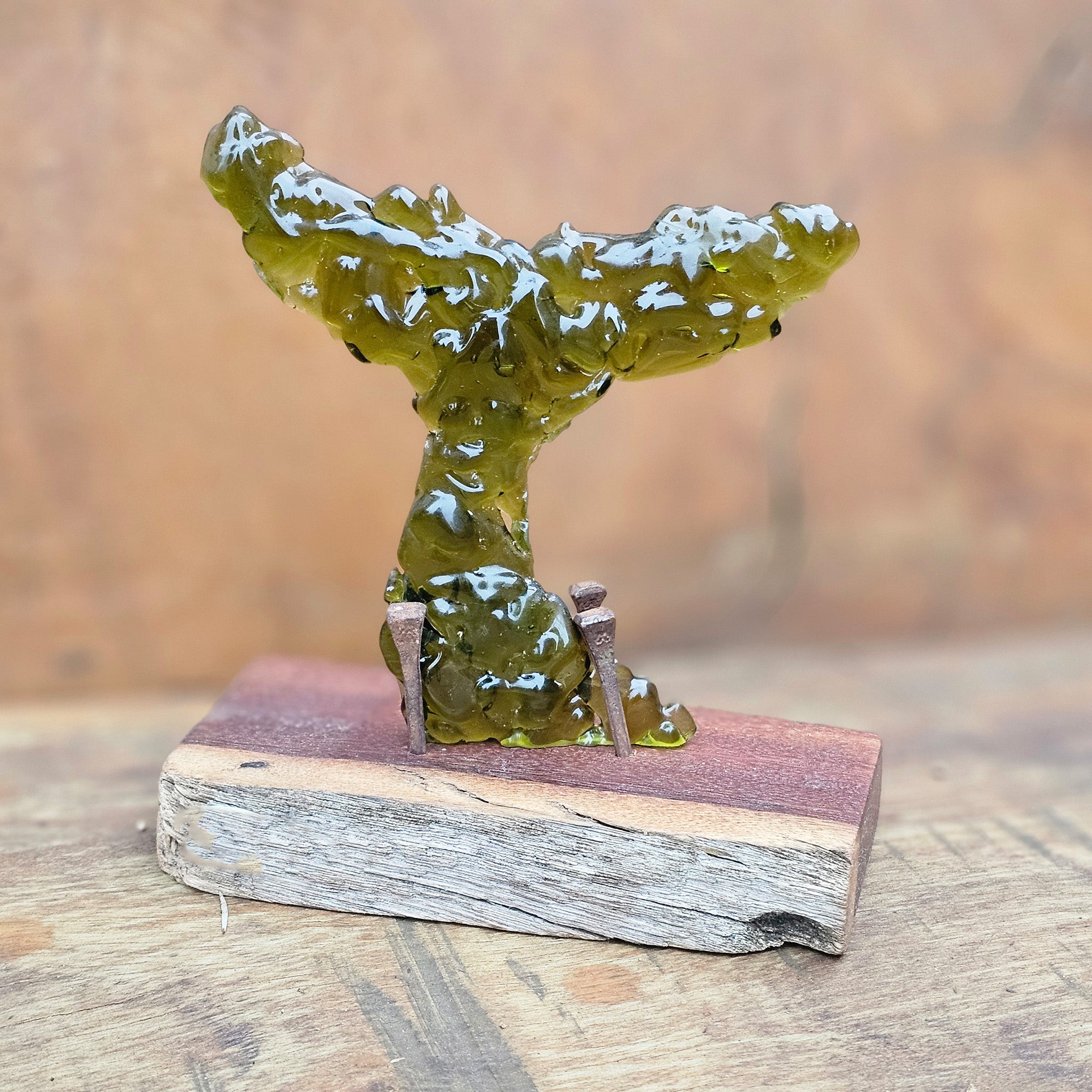 Decorative whale tail sculpture, formed from kelp-green-olive fused glass fragments on a wooden base, with aged rusted nails, against a blurred wooden background.