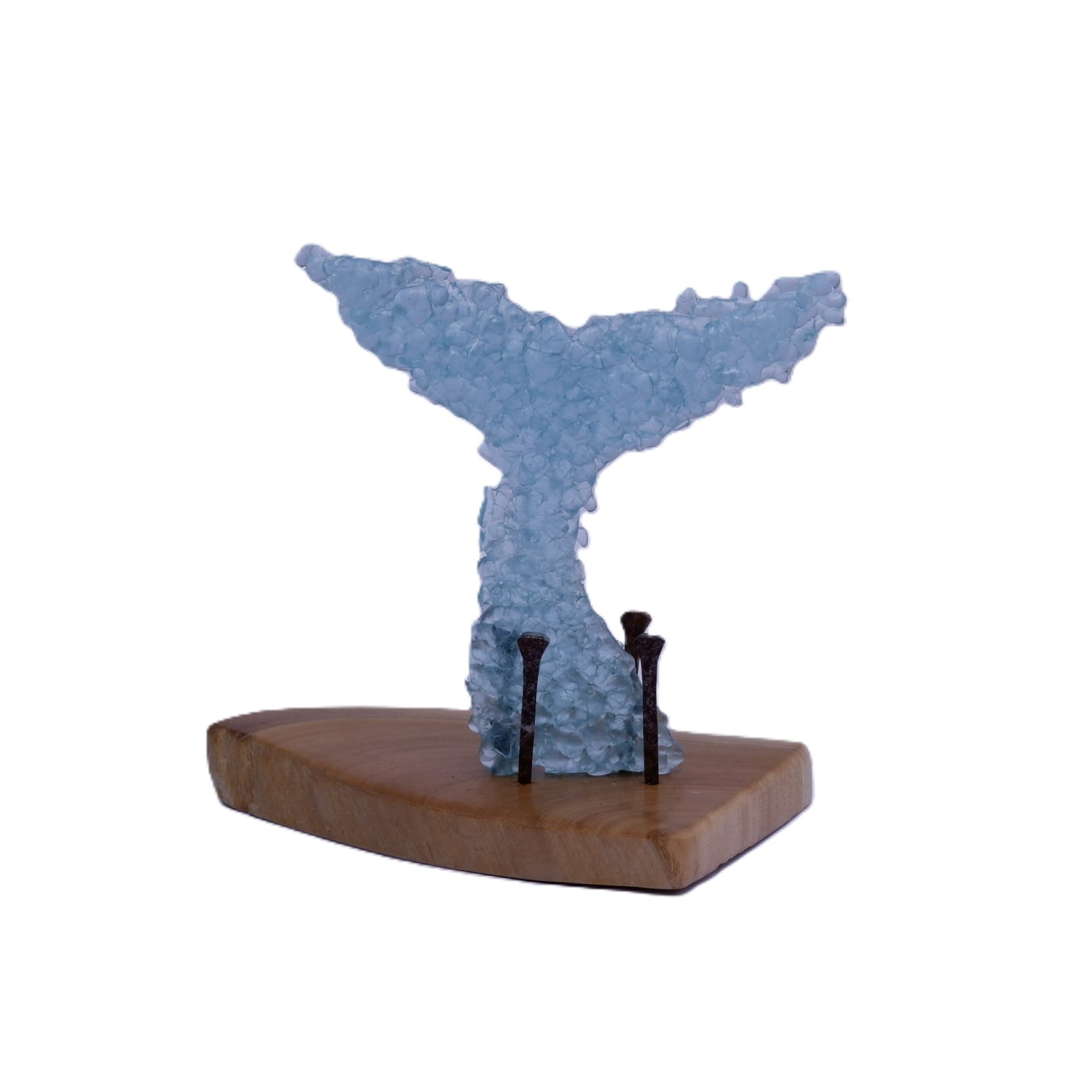 A whale tail made of kiln formed light blue glass fragments displayed on a wooden base supported with 3 visible aged and rusted nails, photographed against a plain white background.