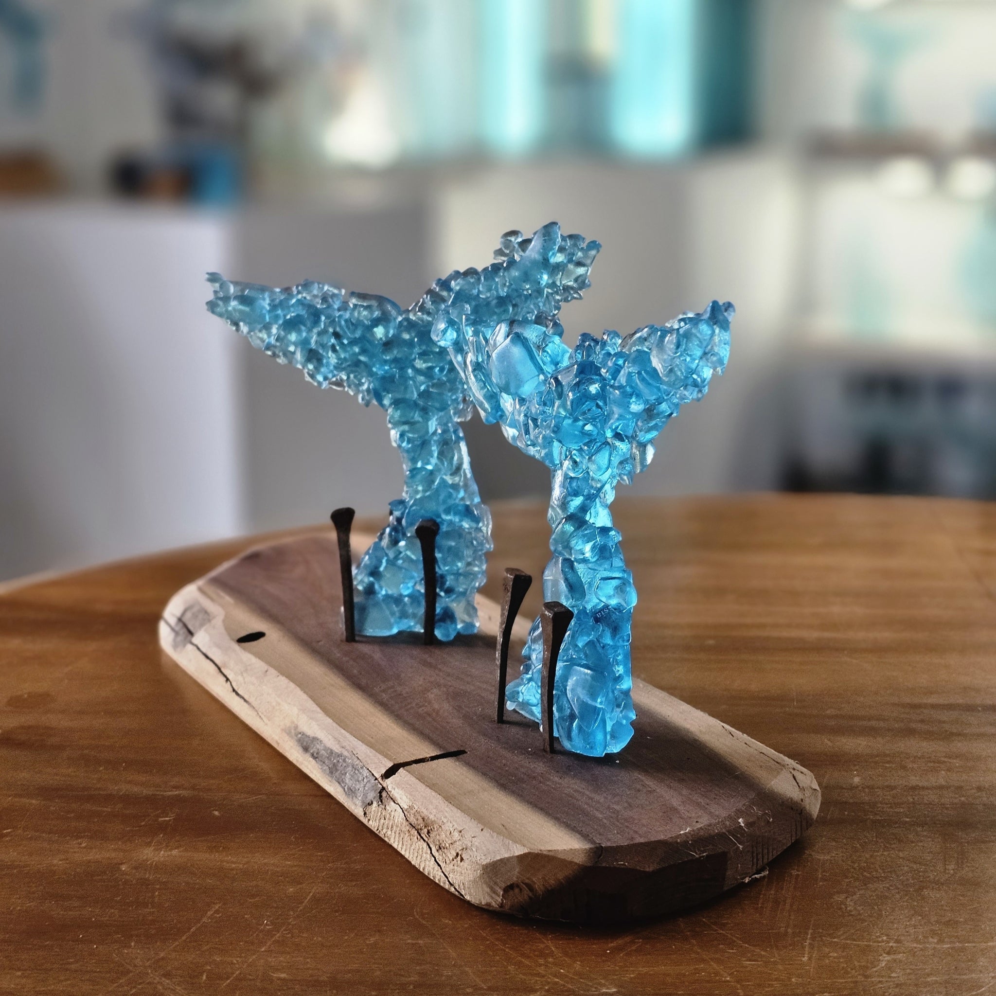 A decorative tabletop sculpture with 2 baby whale tails made from translucent blue fused glass fragments mounted on a rustic live-edge wooden board with aged rusted horse shoe nails, shown on a wooden table in a softly blurred indoor setting.
