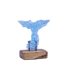 A pale blue whale tail sculpture mounted on a natural wood base with two small aged nails, photographed against a plain white background.