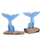 Two decorative whale tail sculptures in translucent pale blue fused glass fragments, mounted on natural wood bases with aged metal nails, shown on a plain white background.