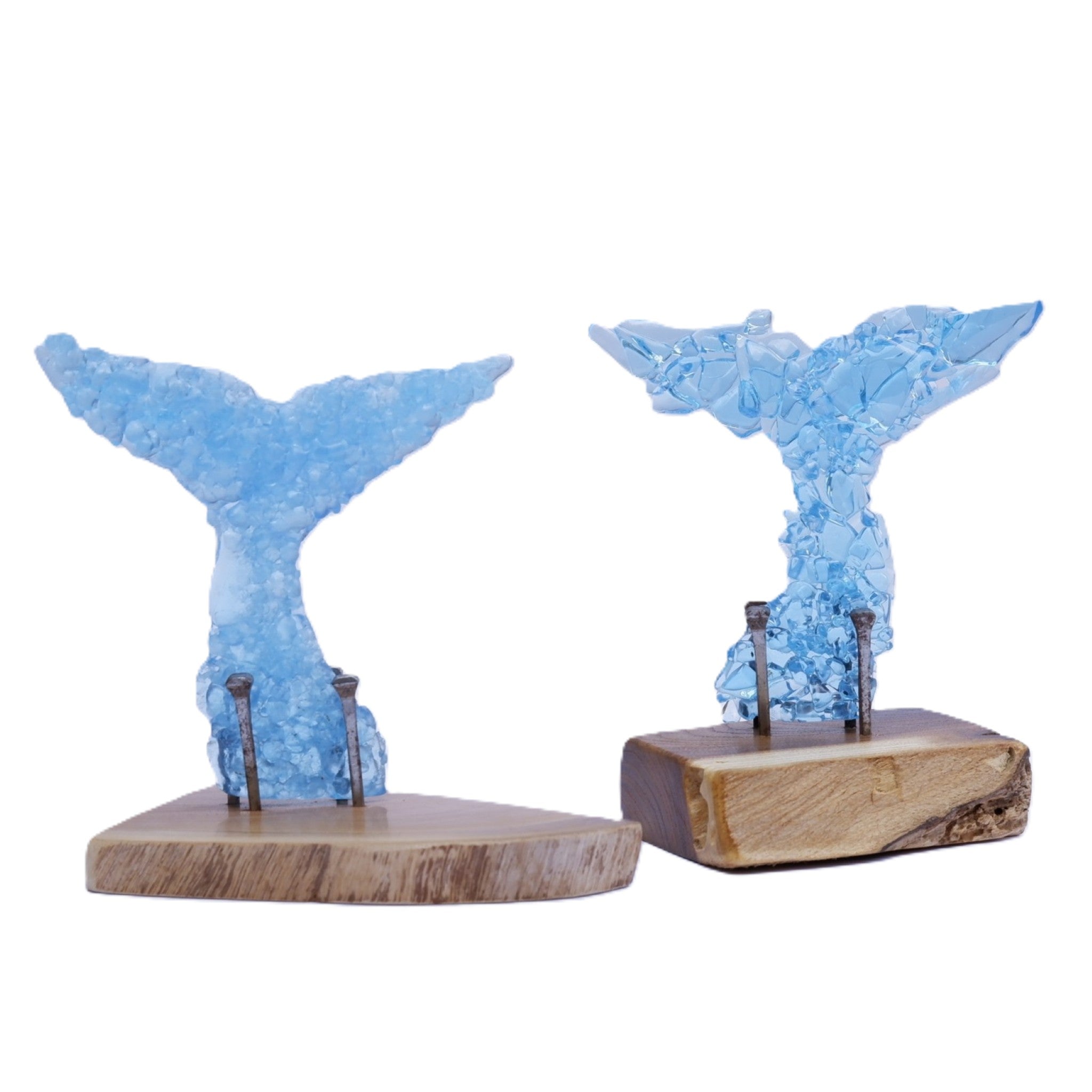 Two decorative whale tail sculptures in translucent pale blue fused glass fragments, mounted on natural wood bases with aged metal nails, shown on a plain white background.