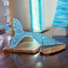 A translucent light blue glass whale tail sculpture resting on a split oiled Australian bush timber base, photographed on a dark wooden table with a softly blurred indoor background.