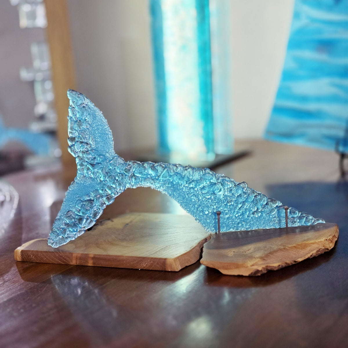 A translucent light blue glass whale tail sculpture resting on a split oiled Australian bush timber base, photographed on a dark wooden table with a softly blurred indoor background.