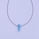 A delicate black stainless steel thread necklace featuring a single polished light blue teardrop styled glass pendant, displayed against a solid light colouredbackground.