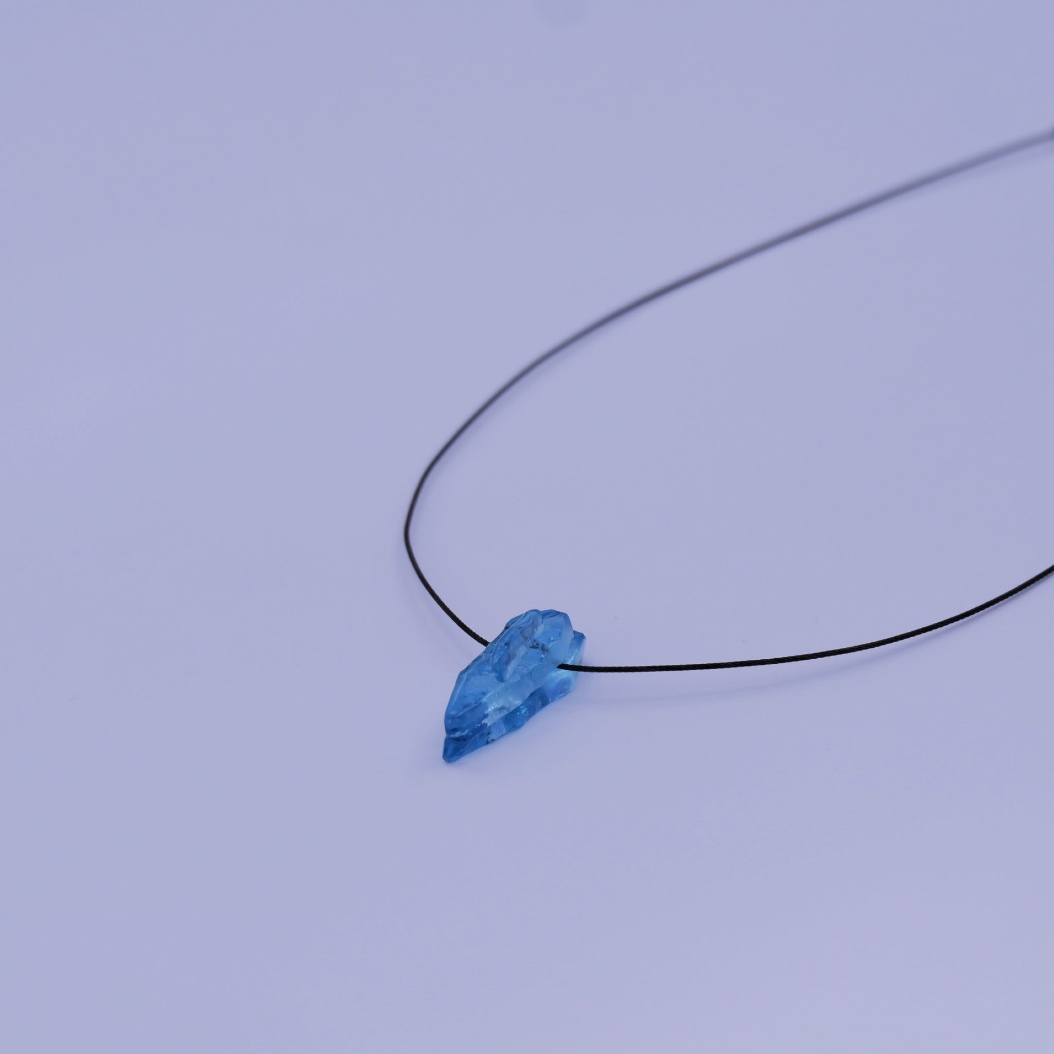 A delicate black stainless steel thread necklace featuring an organic blue teardrop styled glass pendant, photographed against a solid light coloured background.