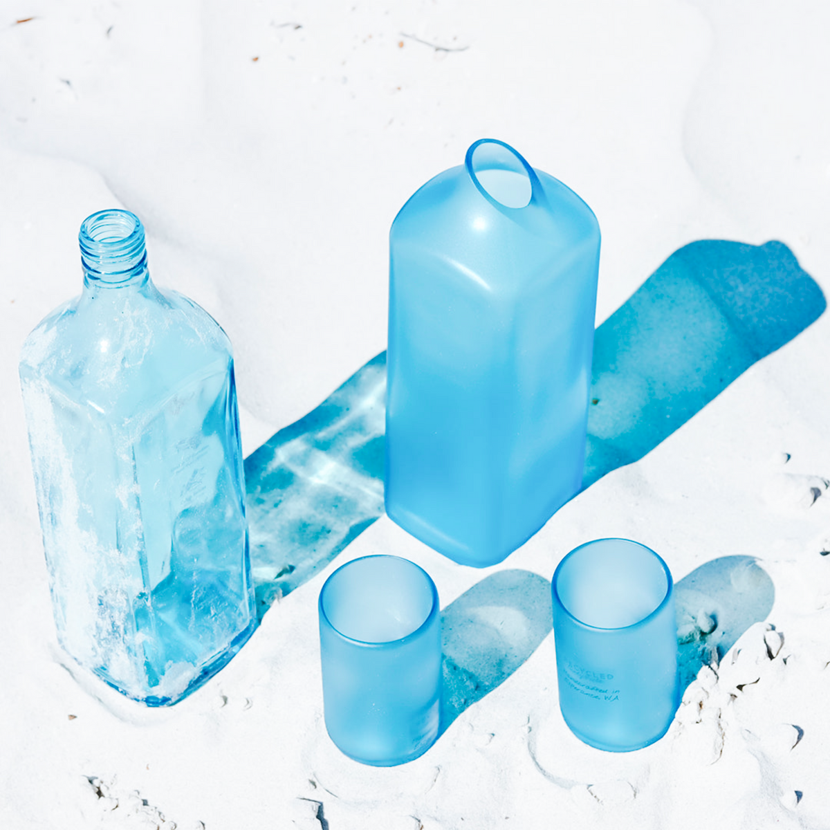 Three translucent light blue glassware pieces arranged on a white surface: a tall rectangular bottle, a matching carafe, and two short tumblers, with blue shadows behind them.