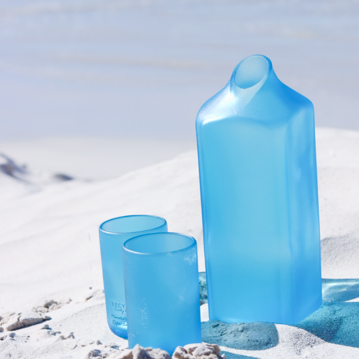 A translucent light blue drinking carafe and two matching round glass tumblers are arranged on white sand with a softly blurred beach or ocean background.