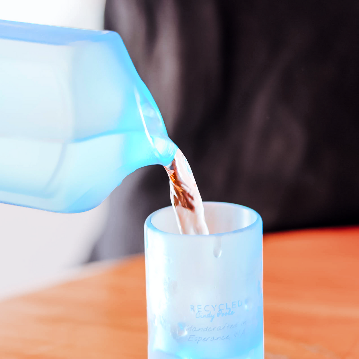 A translucent light blue glass tumbler with branding printed on the front, being filled with water from a matching blue glass carafe. The background is a soft-focus indoor setting with a dark garment and a wooden table.