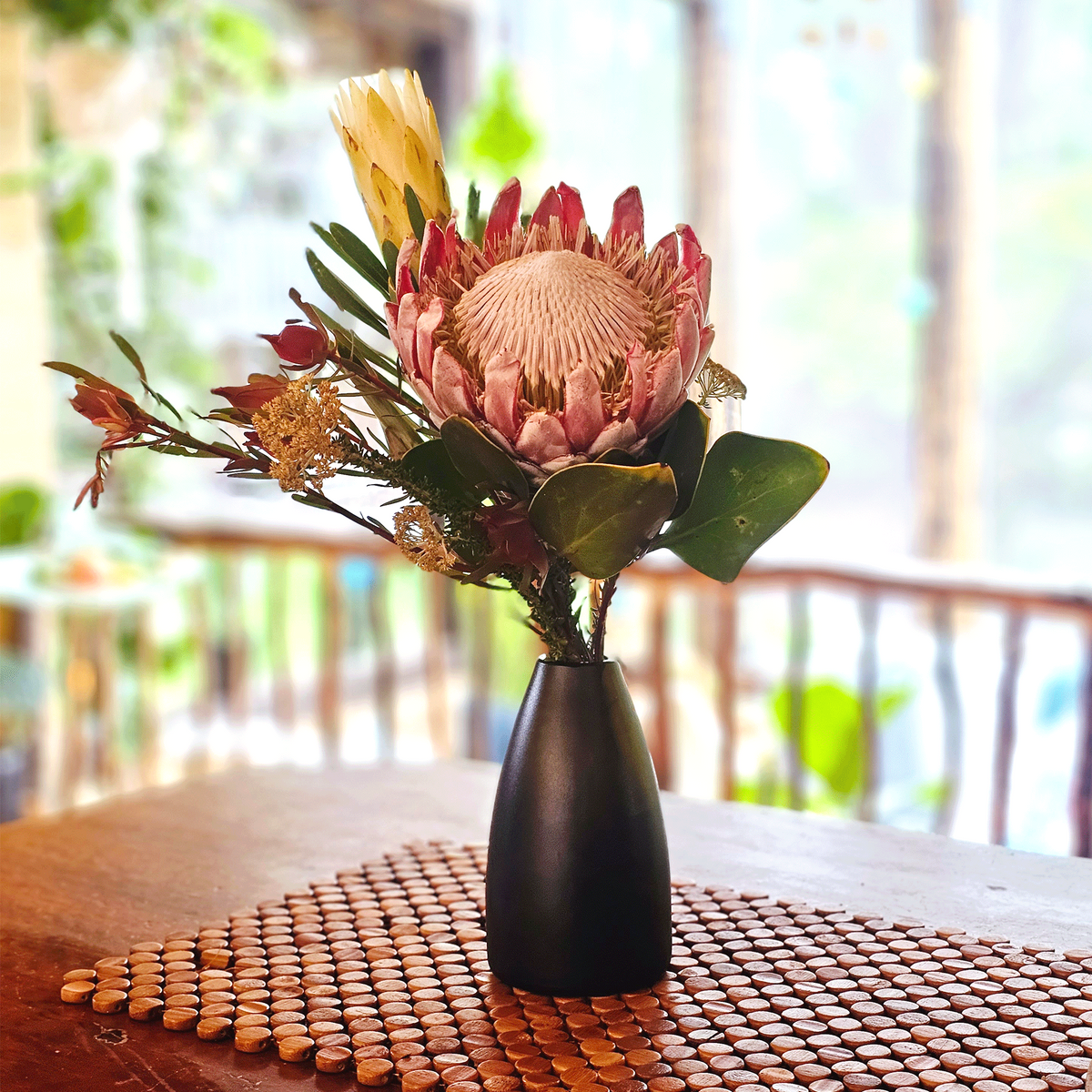 A dark obsidian hued glass vase holding a dried protea flower arrangement with pink, cream, and green botanical elements, set on a woven brown placemat on a wooden table with a softly blurred indoor outdoor background.