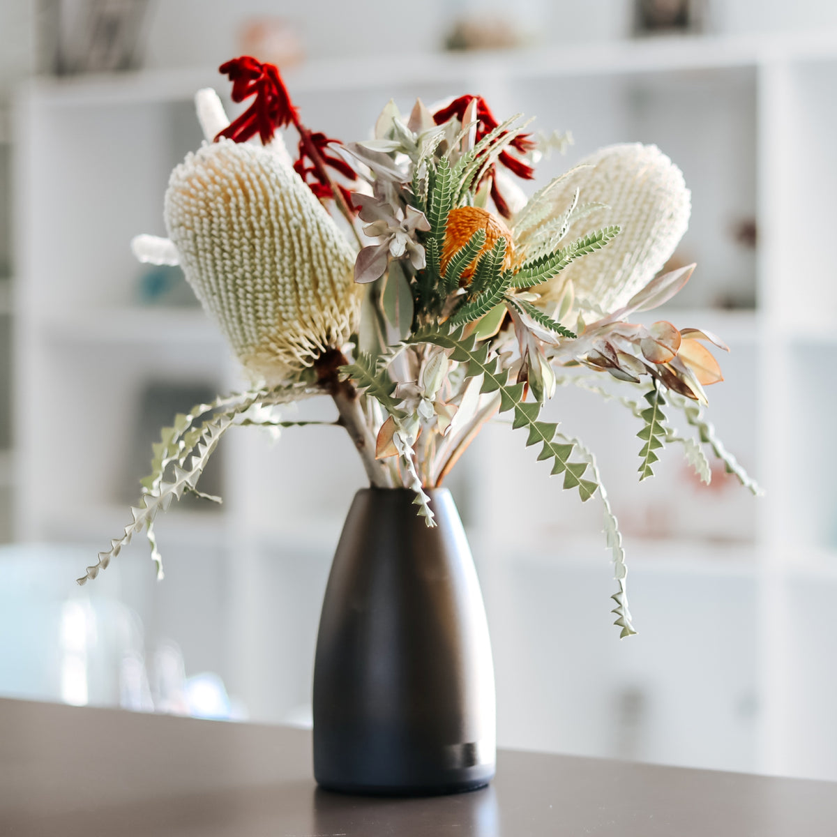 An Australian bush flower arrangement featuring cream banksia blooms, a red kangaroo paw accent, green banksia textured leaves, and small orange flowers in a matte obsidian glass vase, set on a tabletop with a softly blurred indoor background.