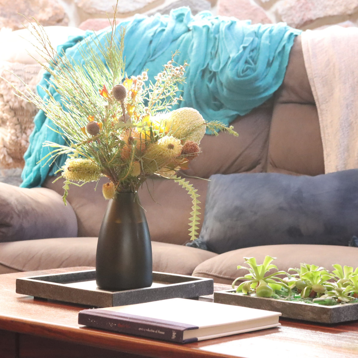 A dark olive green hued glass vase filled with a dried floral arrangement, styled on a wooden coffee table in a cozy living room setting with a brown sofa, blue throw blanket, and small succulent planter.
