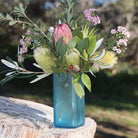 A blue glass vase filled with a australian wildflower arrangement featuring a pink protea, green leaves, yellow banksia, and small pink flowers, set outdoors on a tree stump with a blurred natural background.