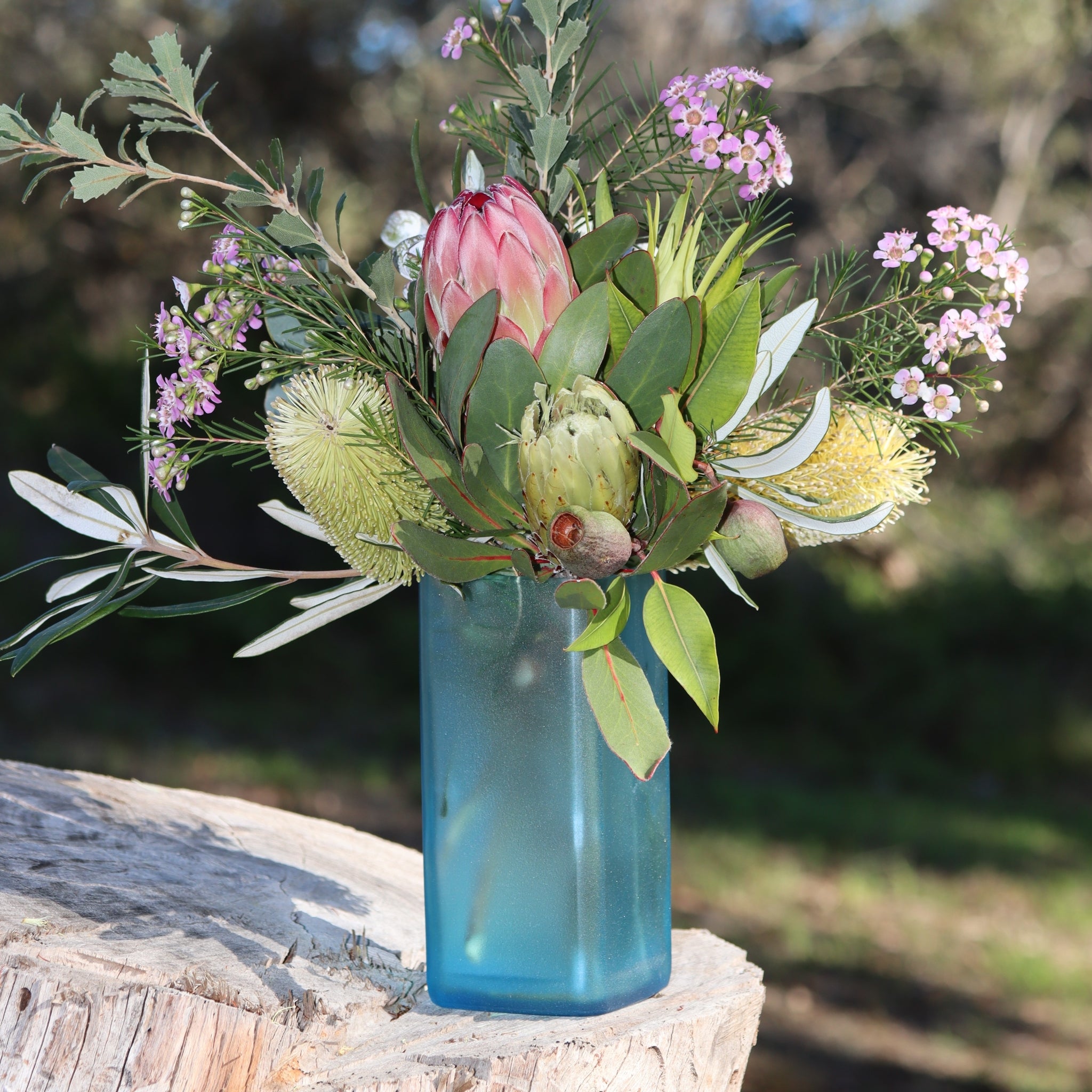 A blue glass vase filled with a australian wildflower arrangement featuring a pink protea, green leaves, yellow banksia, and small pink flowers, set outdoors on a tree stump with a blurred natural background.
