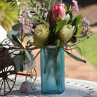 A blue glass vase filled with an Australian native floral arrangement of pink protea, yellow banksia, and small pink flowers, displayed on a white table outdoors.