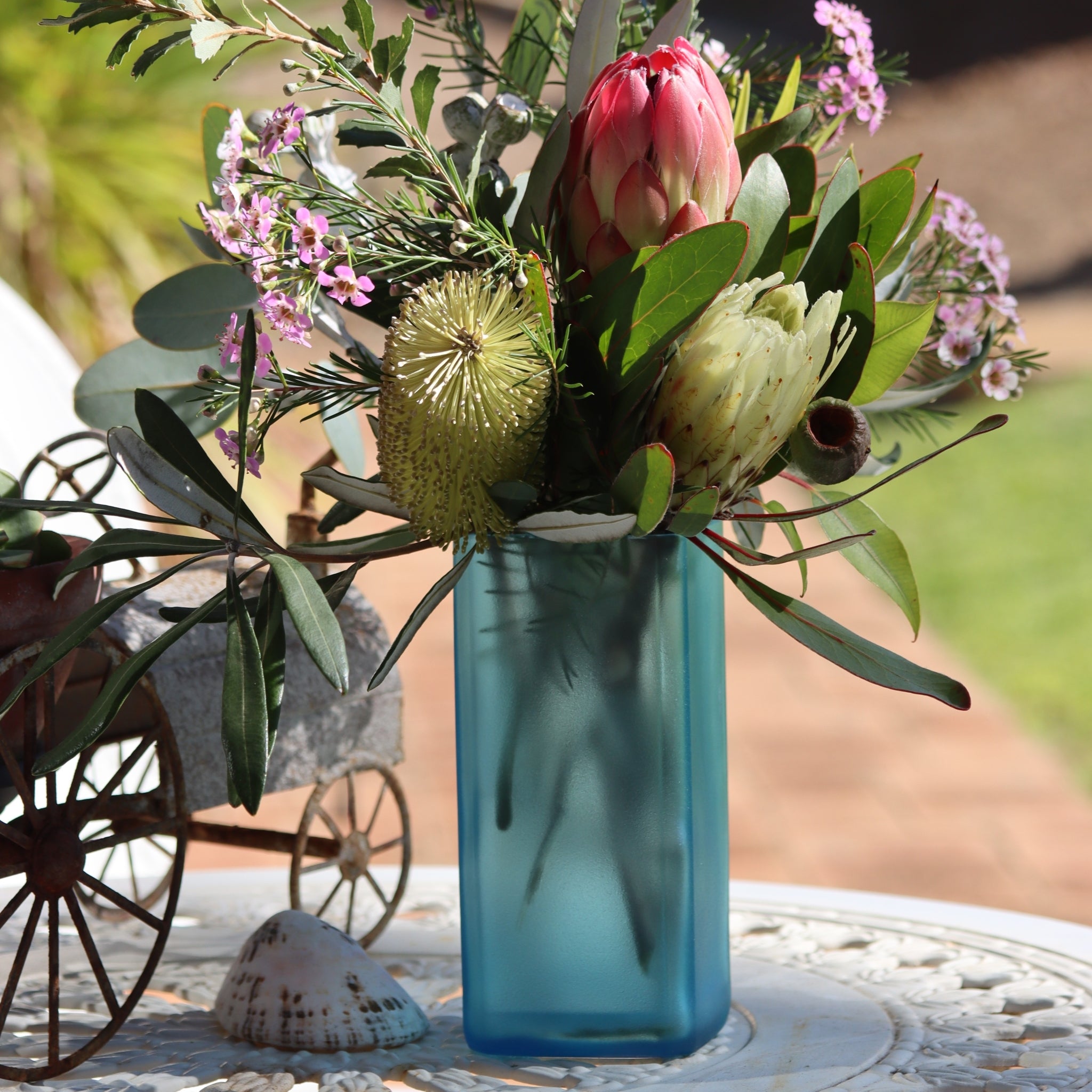 A blue glass vase filled with an Australian native floral arrangement of pink protea, yellow banksia, and small pink flowers, displayed on a white table outdoors.