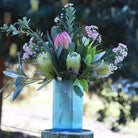 A vibrant blue glass vase filled with a lush Australian-style wildflower bouquet, including pink protea, yellow banksia, and small pink flowers, displayed on a wooden fence post against a soft-focus outdoor background.