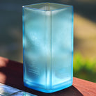 A translucent light blue square glass vase with sateen texture, standing on a wooden surface outdoors with blurred greenery in the background.