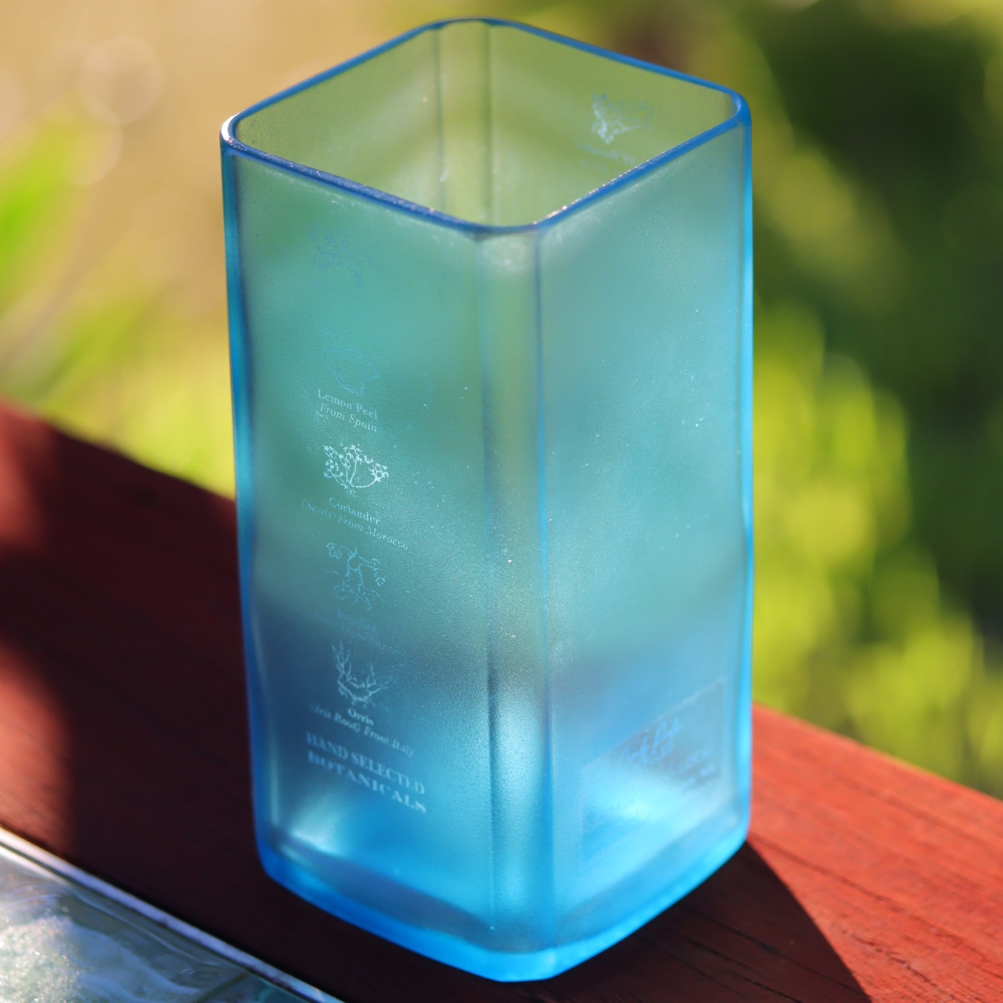 A translucent blue square glass vase with sateen finish and white printed text, photographed outdoors on a wooden surface with blurred green foliage in the background.