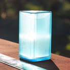A translucent light blue square glass vase brightly lit with sunshine, sitting on a wooden table outdoors with a blurred natural background.