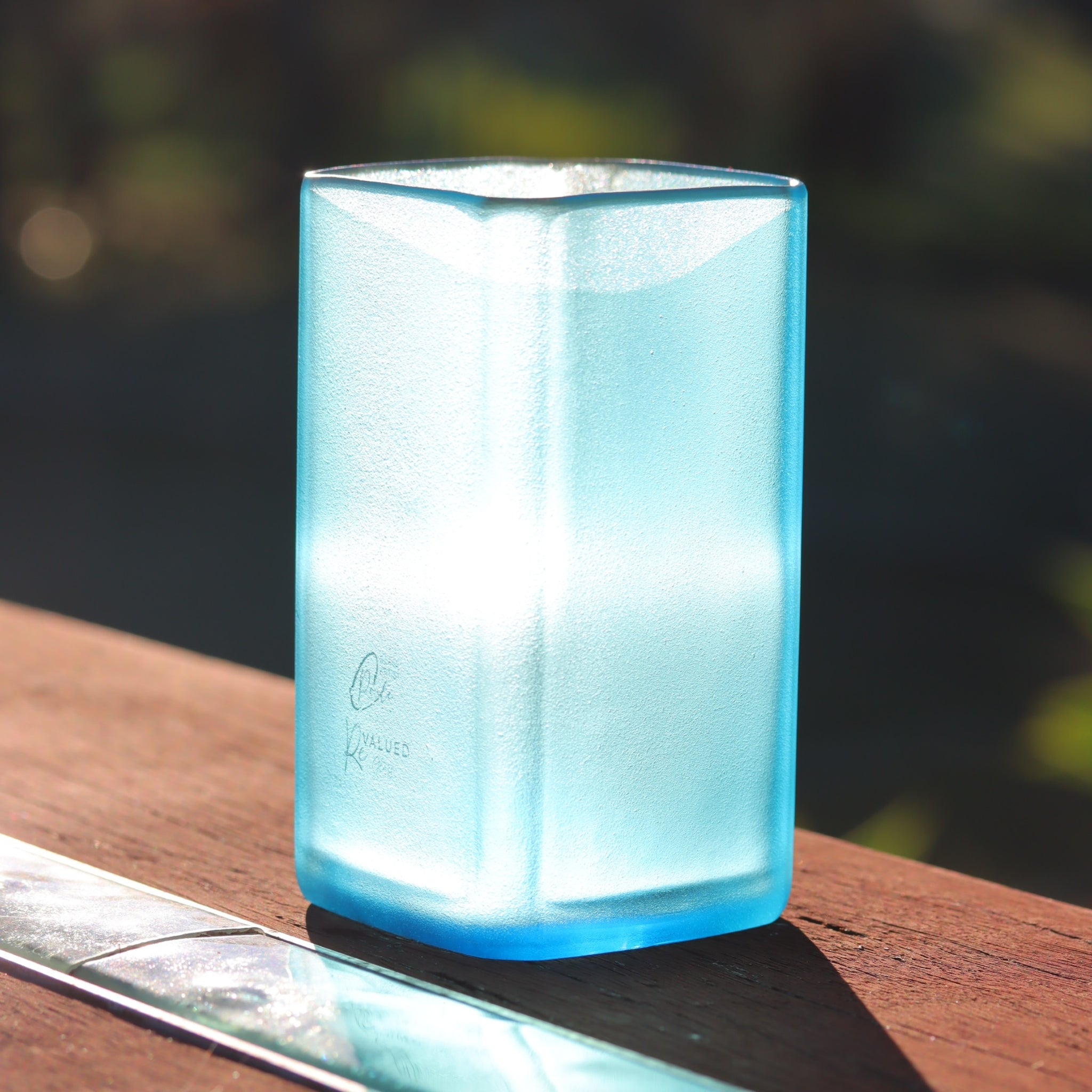 A translucent light blue square glass vase brightly lit with sunshine, sitting on a wooden table outdoors with a blurred natural background.